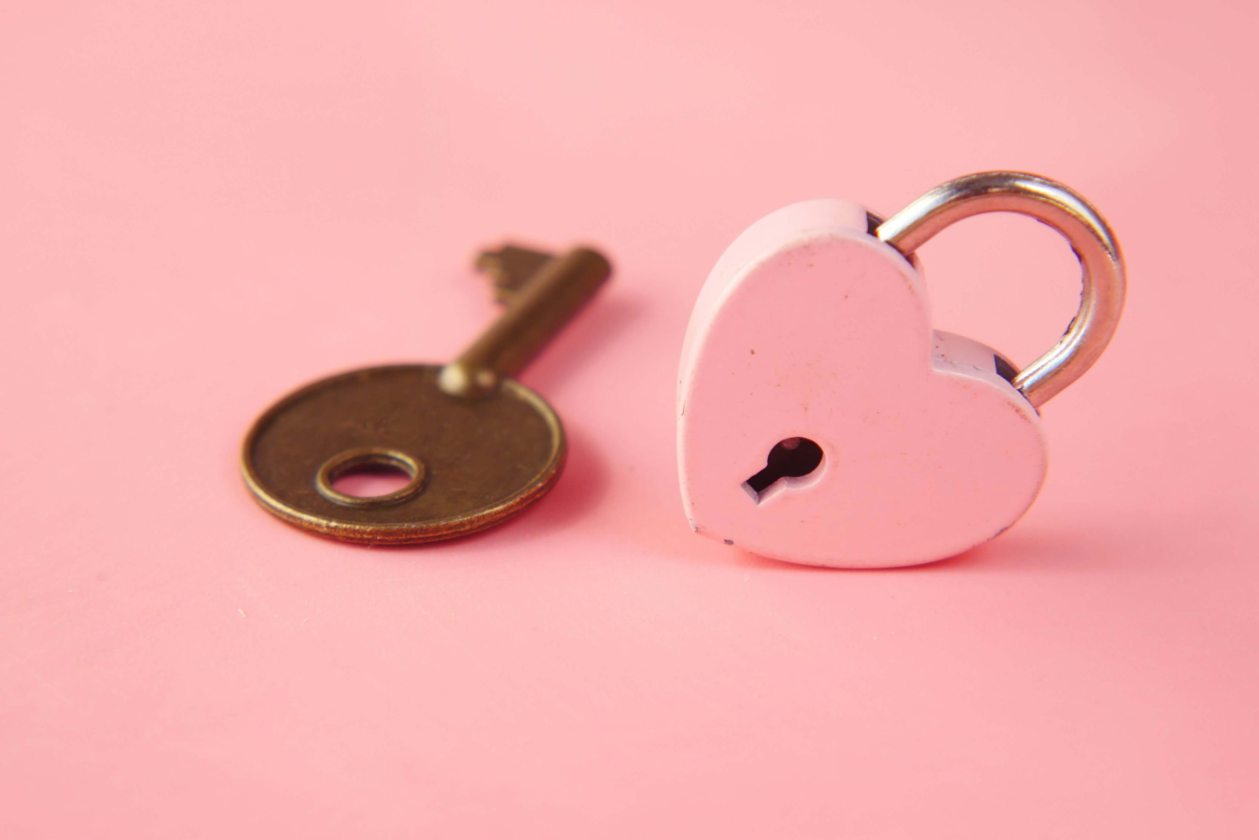 HeartLockKeyTowfiquBarbhuiyaOct2021Large Pink Heart-Shaped Padlock, Titled on its Edge, With Bronze Key Next To It, Against Pink Background
